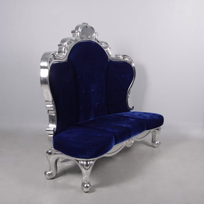 The Grand Deluxe Throne, a three-seat throne in blue tufted velvet with a carved silver frame, photographed at an angle in the merchant's studio.